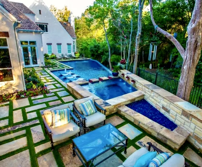 Geometric pool and raised spa in a wooded yard, with in-water loungers, a linear fire pit, and a flagstone-and-turf patio