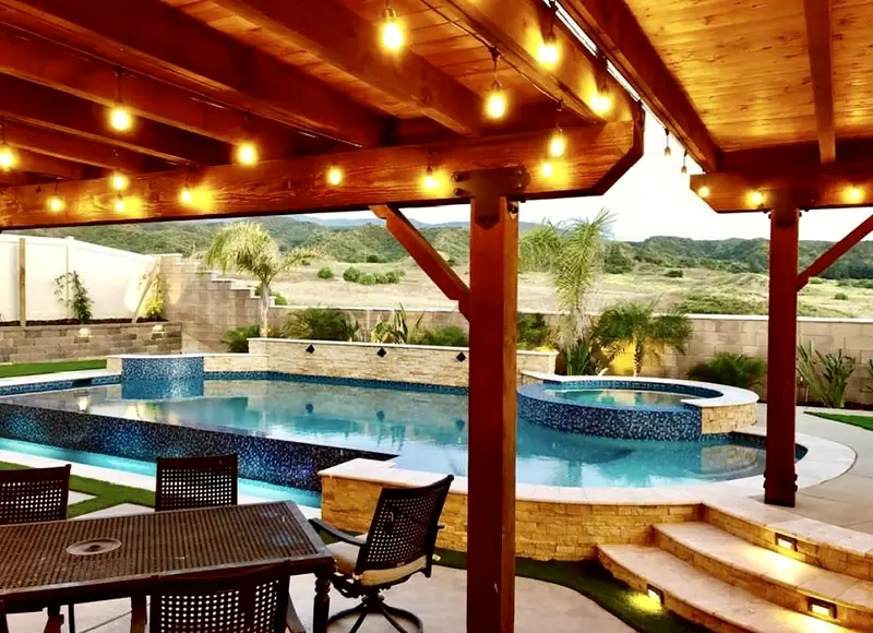 Vanishing-edge pool and raised spa seen from under a string-lit wood pergola at dusk, with hills beyond