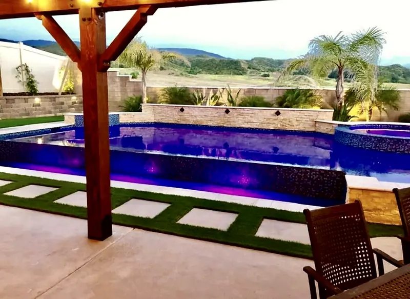 The same pool at night: color LED lighting, a glass-tile vanishing edge, and turf-and-paver stepping stones