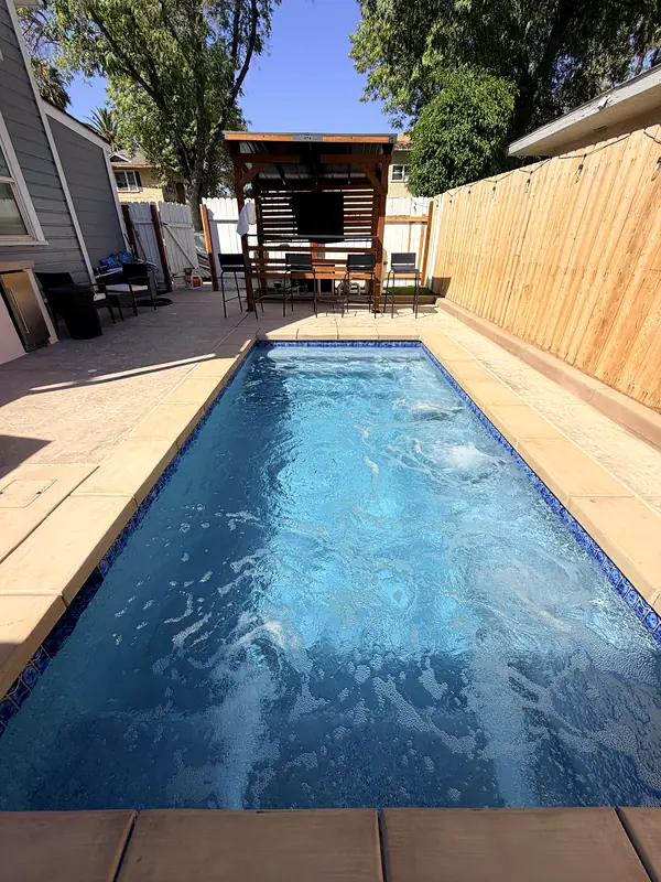 Long, narrow pool filling a side yard, with a glass-tile waterline and a pergola lounge at the far end